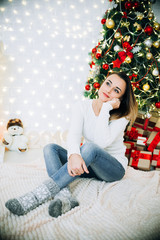 girl in white sweater and jeans sitting on a Christmas tree background, tender and cute