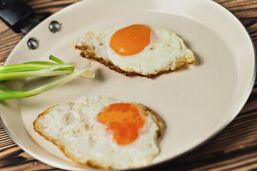 Two fried eggs in metal beige pan and green fresh onion on old wooden brown table