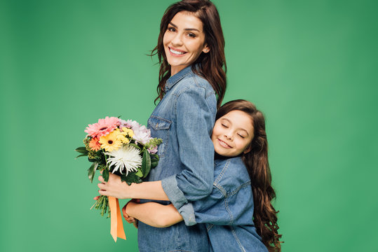 adorable daughter hugging smiling mother with flowers isolated on green