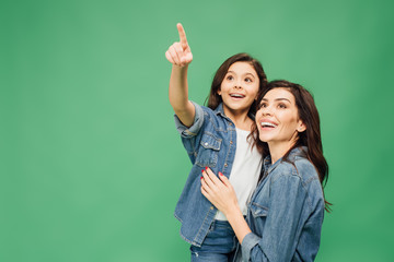 excited daughter pointing with finger while happy mother looking up isolated on green
