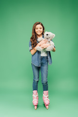 child in roller blades holding teddy bear and looking at camera on green background