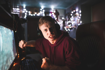 Angry young gamer in headphones plays video games at home on a computer, looks into the camera and gets angry.Emotional teenager sitting at home in a computer room, and an angry loser in the game.