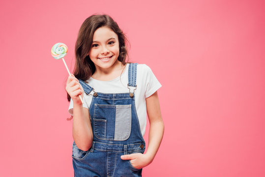 Cheerful Kid Holding Lollipop With Hand In Pocket Isolated On Pink