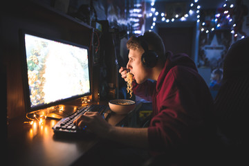 Portrait of a focused gamer playing video games at home on a computer and eats a noodle soup. Young man eats noodles and uses a computer at home. © bodnarphoto