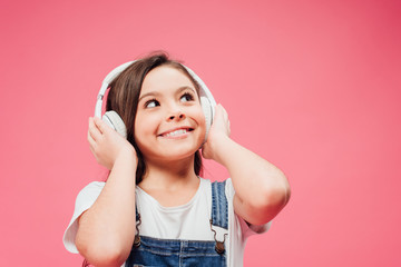 cheerful child listening music and touching headphones isolated on pink
