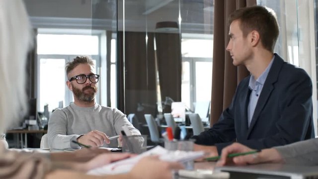 Medium shot of young bearded businessman in glasses sitting at office desk and talking to group of colleagues during meeting