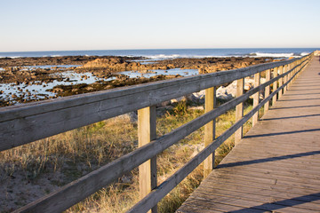 Obraz premium Wooden pier with grass on Atlantic ocean coast, Portugal. Wooden boardwalk to sea with rocks on shore. Empty walkway in dunes at dawn. Perspective background. Tropical travel concept. Peaceful place.