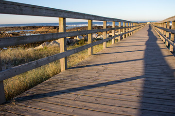 Obraz premium Wooden pier with grass on Atlantic ocean coast, Portugal. Wooden boardwalk to sea with rocks on shore. Empty walkway in dunes at dawn. Perspective background. Tropical travel concept. Peaceful place.