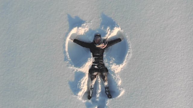 Top aerial view of young happy smiling girl making by arms snow angel figure and lying in snow, winter outdoor activity