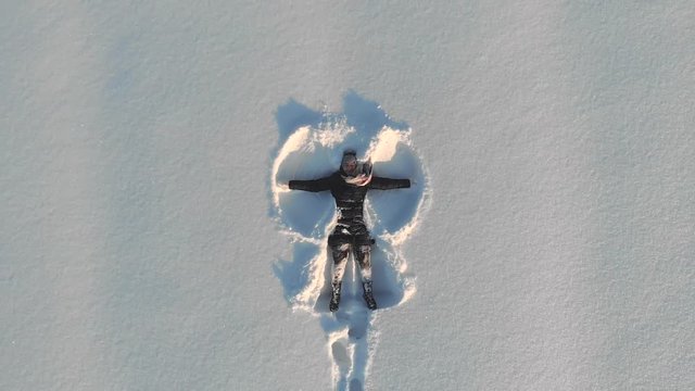 Top aerial view of young happy smiling girl making by arms snow angel figure and lying in snow, winter outdoor activity