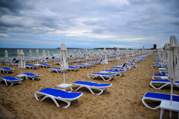 empty empty beach with folded beach umbrellas, blue beach chairs and a stormy sky 