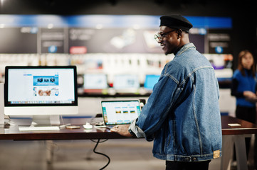 Stylish casual african american man at jeans jacket and black beret using new laptop at electronics...