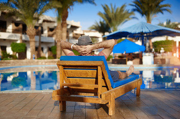 Young man in hat lies on louger near swimming pool for relax summer vacation concept