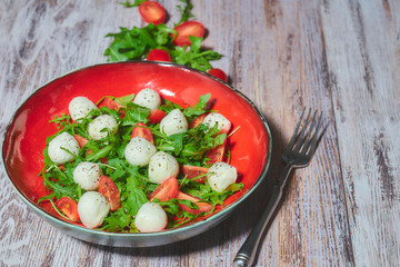 a plate of fresh salad with arugula, cherry tomatoes, mozzarella cheese on a white, shabby, wooden table. vertical view. Copy space.