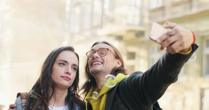 Portrait of the young good looking boyfriend and girlfriend in stylish looks laughing, kissing and taking selfie photos on the smartphone camera. Outdoor. Close up.