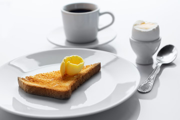 Soft boiled egg, toasted bread, butter,  cup of coffee and spoon on white table. Selective focus.