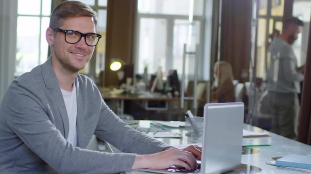 Tracking medium shot of cheerful young Caucasian man in glasses sitting at office desk, typing on laptop computer and smiling at camera