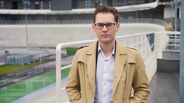 Closeup portrait of attractive caucasian man looking straight at camera with neutral facial expression outdoors hands in the pockets