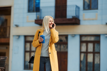 Fototapeta premium A young blond woman in a yellow bright coat is standing in the middle of the street and talking on the phone. Waiting for a meeting of friends. Holds a cup of coffee to go.