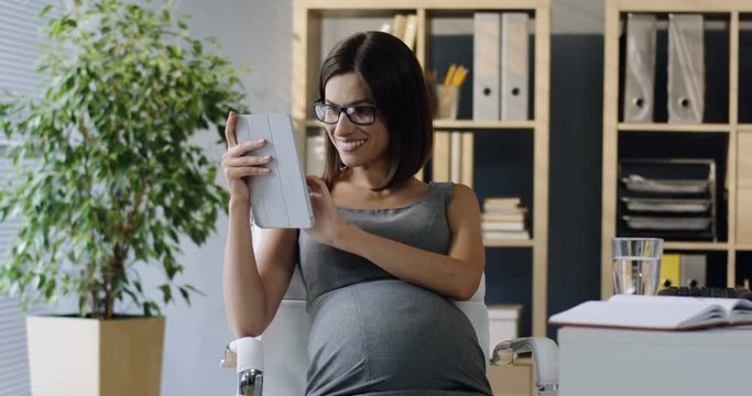 Good looking Caucasian pregnant woman sitting at her workplace in the office and having a videochat on the tablet device.