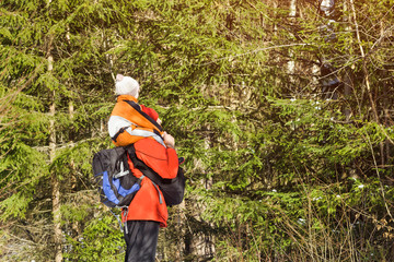 Man with backpack and son on shoulders stands against the background of coniferous trees in the forest. Winter day. Side view. Close-up