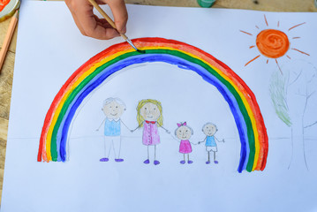 Happy family concept. Child draws on a sheet of paper: father, mother, boy and girl hold hands against background of rainbow and sunny sky. Close-up.