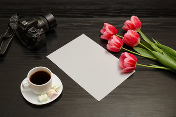 Fototapeta na wymiar Holiday concept. Bouquet of pink tulips, a cup of coffee, dslr camera and sheet of paper on a black wooden background