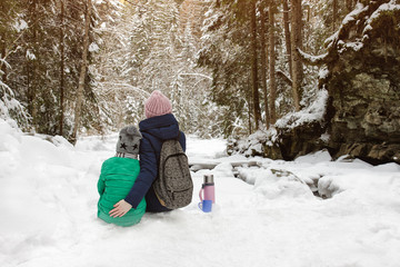 Fototapeta premium Mom with her son sitting and embracing in the background of snow-covered forest. Winter cloudy day. Back view