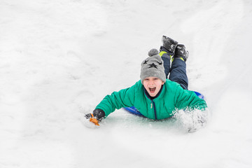 Fototapeta na wymiar Laughing boy rides on his stomach from a hill on snow saucer. Seasonal concept. Winter day