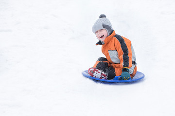 Fototapeta na wymiar Happy laughing small boy slides down the hill on snow saucer. Seasonal concept. Winter day