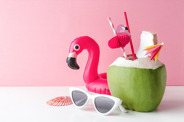 Fresh coconut on a pastel pink background with flamingo inflatable drink holder and sunglasses,...