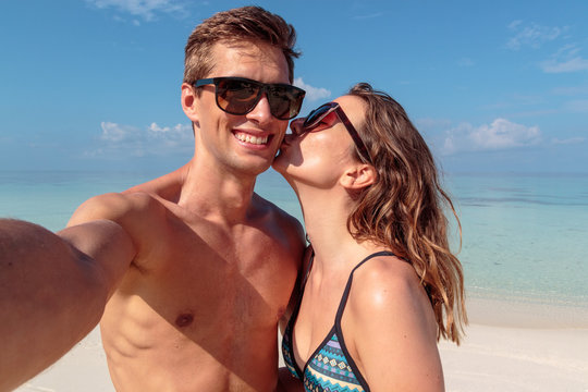 happy young couple taking a selfie, clear blue water as background. Girl kissing his boyfriend