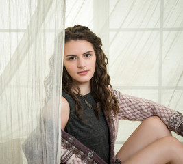 Beautiful, young woman in sweater sitting in front of draped, white window