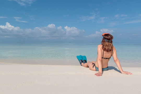 picture from back of a young woman with flippers and mask seated on a white beach in the Maldives. Crystal clear blue water as background