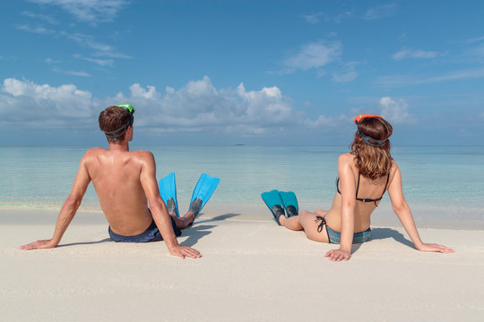 picture from back of a young couple with flippers and mask seated on a white beach in the Maldives. Crystal clear blue water as background