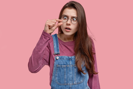 Young woman shows small amount of something, demonstrates unimpressive size, shapes tiny thing, has scared expression, dressed in stylish clothes, models against pink studio wall. Little object
