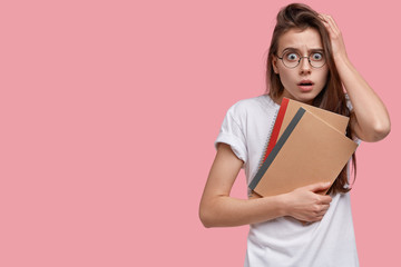 Shocked Caucasian schoolgirl stares with bugged eyes, keeps hand on head, notices something incredible, dressed casually, holds notepads, preapares for examination session, isolated on pink wall