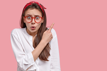 Worried European woman has fearful expression, points at upper right corner, wears white shirt,...