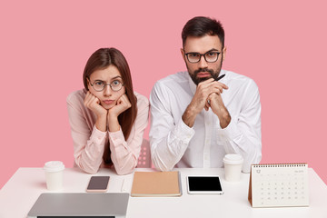 Serious young coworkers think on common solution, look directly at camera, dressed in stylish clothes, hold chin, have gadgets and stationary arranged in order on table, pose over pink background