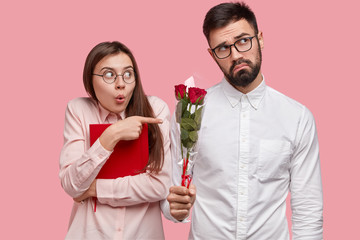 Waist up shot of shocked European woman points at groupmate who has sad expression, holds red roses, wants to congratulate her with successfully passed exam. Young couple study and have date