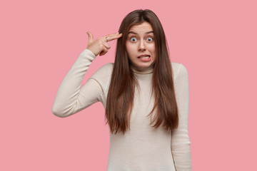 Fototapeta premium Photo of nervous puzzled Caucasain lady makes suicide gesture in temple, shoots in head, has displeased facial expression, wears casual white sweater, isolated over pink background. Body language