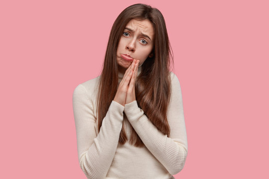 Desperate sorrowful young charming woman with long hair, keeps palms in praying gesture, asks God to make dreams come true, beggs for favour, says please help me out, isolated over pink background