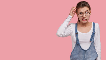 Photo of puzzled woman scratches head in bewilderment, thinks about finding right solution, wears...