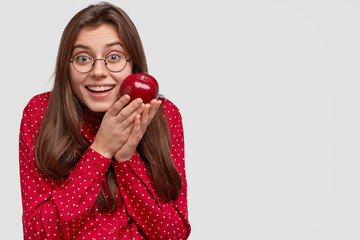 Smiling European woman with pleased expression, carries red apple, dressed in fashionable clothes, round spectacles, enjoys eating fruit, isolated over white background, blank space for your slogan