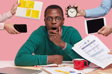 Photo of scared dark skinned guy holds chin, opens mouth widely, wears glasses, surrounded with...
