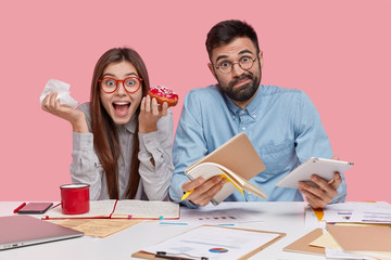 Indoor shot of woman and man coworkers eat tasty doughnut, write down notes in notebook, use modern technologies, collaborate for making financial report with handsome man, isolated on pink.