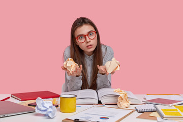 Emotional female nerd has shocked gaze directly at camera, stares through spectacles, holds crumpled paper in both hands, writes financial report, cramms material for exam, isolated on pink wall