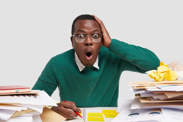 Photo of astonished black man keeps hand on head, jaw dropped, wears big spectacles, writes notes in notepad, dressed in green jumper, isolated over white studio background. Reaction concept.