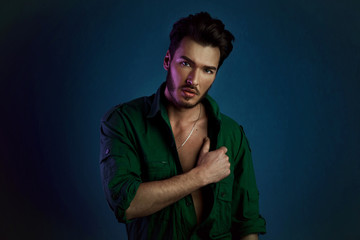 Fashion portrait of young man in green shirt and style haircut poses over dark blue wall with purple contrast color light