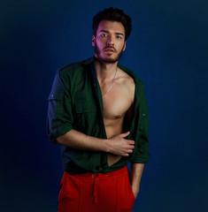 Fototapeta na wymiar Fashion portrait of young man in green shirt and red pants poses over dark blue wall with contrast light